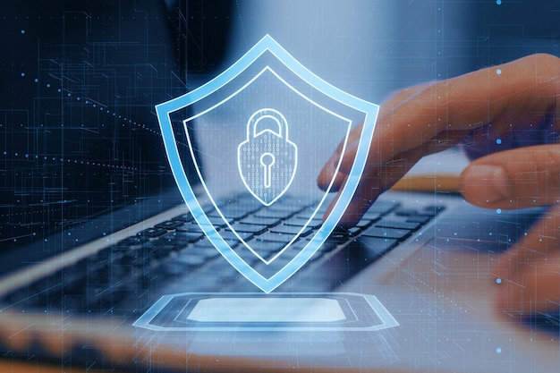 Una ilustración de un escudo de seguridad digital que se superpone a una computadora portátil con una mano del usuario en un fondo de tecnología azul oscuro que simboliza la protección cibernética