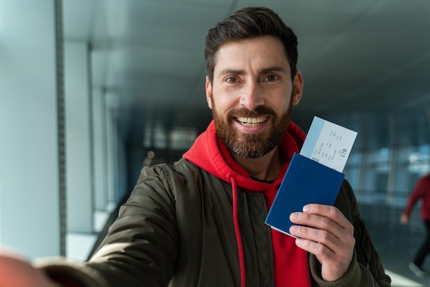 Im bereit für neue Zukunft. Junger kaukasischer überglücklicher mann, der am flughafen mit pass und ticket in der hand in die kamera schaut. Er steht und trägt Gepäck vor dem Flug