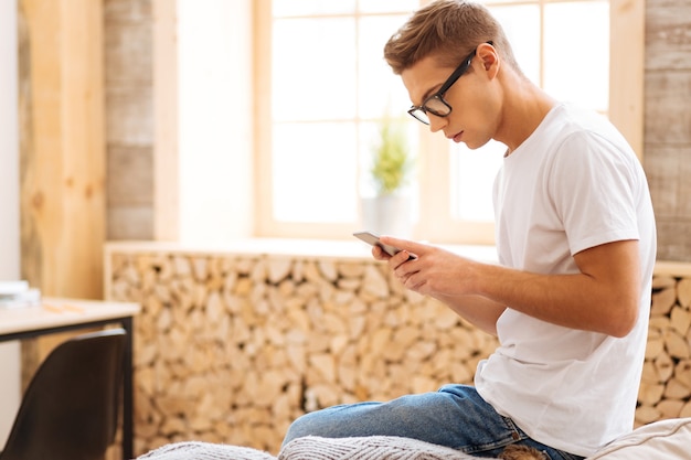 Im Blues. Gut aussehender unglücklicher junger Mann, der eine Brille trägt und sein Telefon benutzt, während er auf dem Sofa sitzt