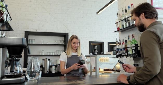 Im Café macht die Frau Kaffee zum Mitnehmen für einen Kunden, der mit kontaktloser Zahlung per Kreditkarte im Café mit digitalem Transaktionsservice Geld ausgibt.