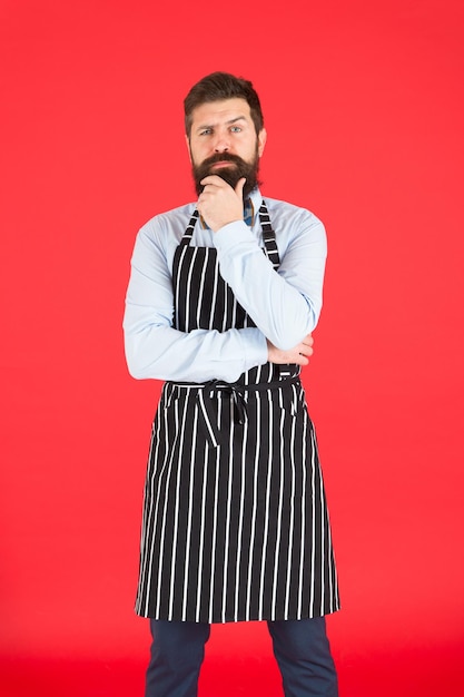 Im Dienst in der Küche Mann mit Bart kochen Hipster-Schürze Hipster-Koch roter Hintergrund Bärtiger Mann Küchenchef Restaurantpersonal und Service Gepflegter Kellner bei der Arbeit Hipster-Café-Konzept