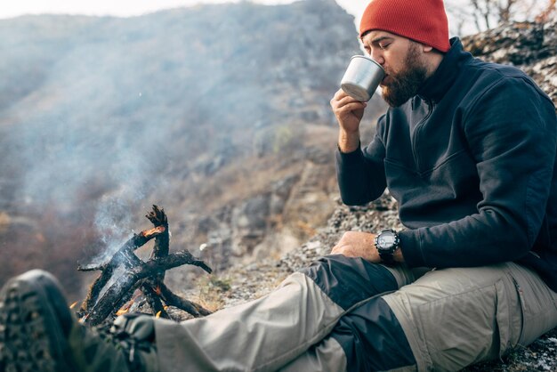 Im Freien Bild des jungen Entdeckermannes, der heiße Getränke in den Bergen trinkt, die in der Nähe des Lagerfeuers sitzen und sich nach dem Trekking entspannen