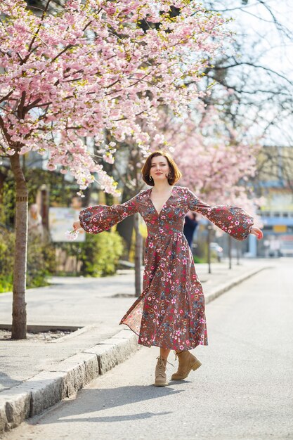 Im Frühling geht eine Frau eine blühende Straße mit Sakura-Bäumen entlang. Ein Mädchen in einem langen, eleganten Vintage-Seidenkleid spaziert zwischen den blühenden Bäumen