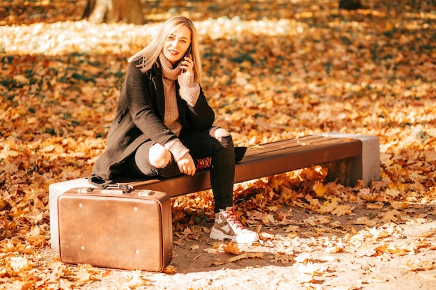 Im Herbstpark der Stadt, zwischen goldenen Laub, sitzt eine attraktive Blondine im schwarzen Mantel auf einer Bank und spricht auf dem Smartphone, neben ihr steht ein alter Koffer. Lebensstil. Leben im Augenblick
