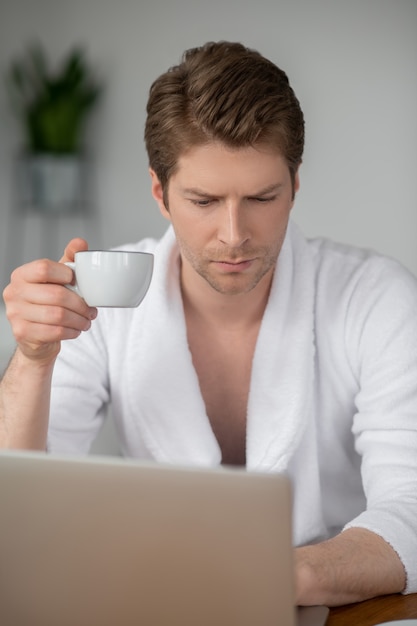 Im Internet. Ein gutaussehender Mann im weißen Bademantel sitzt am Laptop und verbringt Zeit online