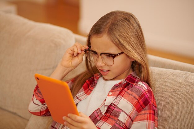 Im Internet. Nettes blondes Mädchen mit einem Gerät in den Händen, das auf dem Sofa sitzt