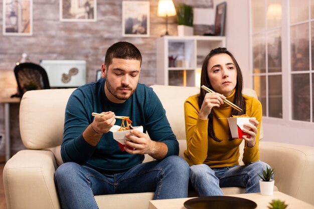 Im modernen gemütlichen wohnzimmer genießt das paar nudeln zum mitnehmen, während sie bequem auf dem sofa fernsehen