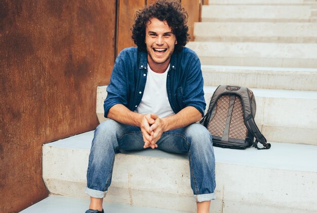 Imagem ao ar livre de estudante jovem sorridente bonito com mochila nas escadas de concreto na rua da cidade Estudante positivo masculino vestindo roupa casual relaxante ao ar livre Pessoas e estilo de vida