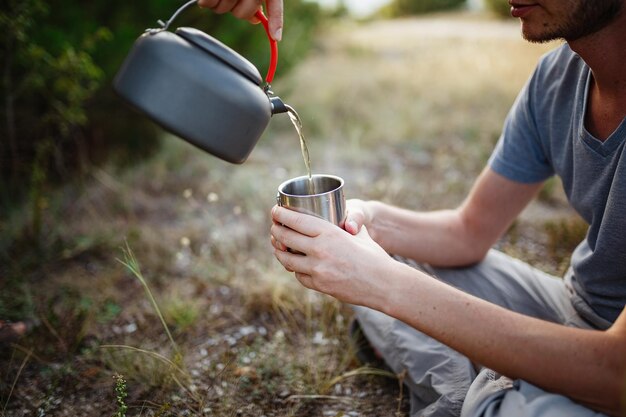Imagem ao ar livre do jovem explorador bebendo bebida quente nas montanhas sentado e relaxando após o trekking