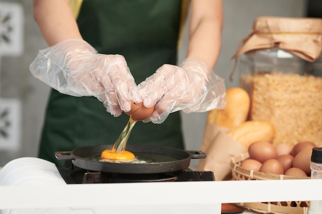 Imagem aproximada de cozinheira usando luvas de proteção enquanto quebra ovos na frigideira