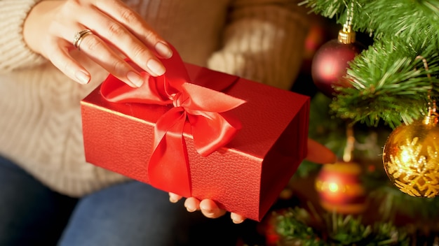 Imagem aproximada de mãos femininas puxando a fita vermelha e abrindo a caixa de presente de Natal da SAnta