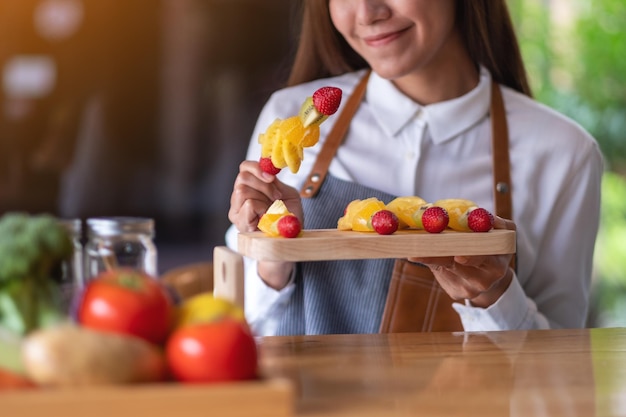 Imagem aproximada de uma bela chef feminina segurando frutas frescas misturadas em espetos em uma placa de madeira