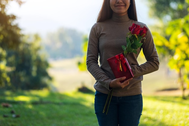 Imagem aproximada de uma bela jovem asiática segurando uma flor de rosas vermelhas e uma caixa de presentes no parque