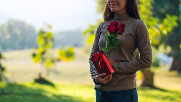 Imagem aproximada de uma bela jovem asiática segurando uma flor de rosas vermelhas e uma caixa de presentes no parque