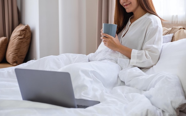 Imagem aproximada de uma bela jovem asiática tomando café enquanto olha para o laptop em uma cama branca e aconchegante em casa