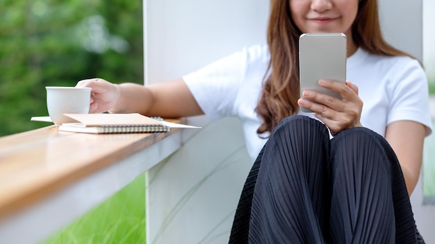 Imagem aproximada de uma bela jovem asiática tomando café enquanto usa o celular ao ar livre