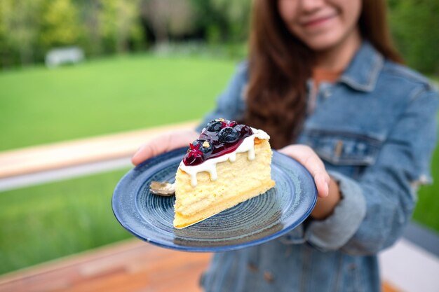 Imagem aproximada de uma jovem asiática segurando e mostrando um prato de cheesecake de mirtilo