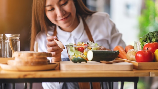 Imagem aproximada de uma jovem chef asiática cozinhando e comendo salada de legumes frescos misturados na cozinha