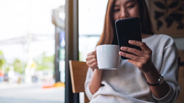 Imagem aproximada de uma linda mulher asiática segurando e usando o celular enquanto bebia café no café