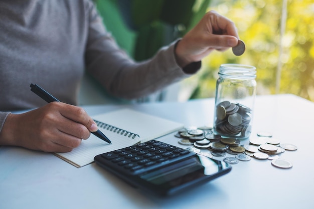 Imagem aproximada de uma mulher colocando moedas em uma jarra de vidro calculando e tomando nota para economizar dinheiro e conceito financeiro