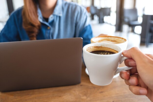 Imagem aproximada de uma mulher e um homem tinindo xícaras de café juntos enquanto trabalhava no computador portátil