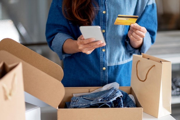 Imagem aproximada de uma mulher usando um telefone celular e um cartão de crédito para fazer compras online com uma sacola de compras e uma caixa postal com roupas na mesa