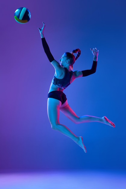 Imagem completa dinâmica de mulher atleta profissional jogando vôlei contra fundo de estúdio azul em luz neon Conceito de anúncio de ação de passatempo de saúde de competição esportiva profissional