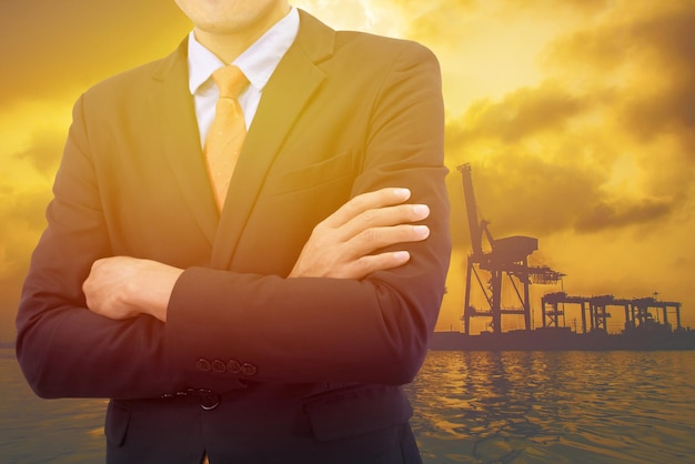 Foto imagem composta digital de um empresário com os braços cruzados de pé contra o porto durante o pôr do sol