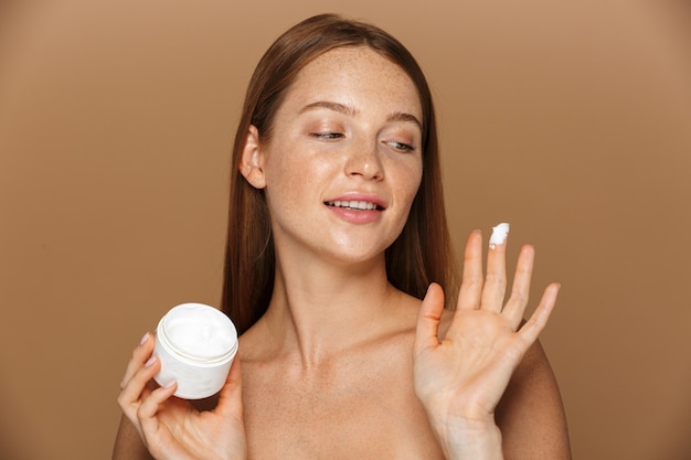 Imagem da beleza de uma mulher sem camisa satisfeita sorrindo e segurando um frasco com creme facial, isolado sobre um fundo bege