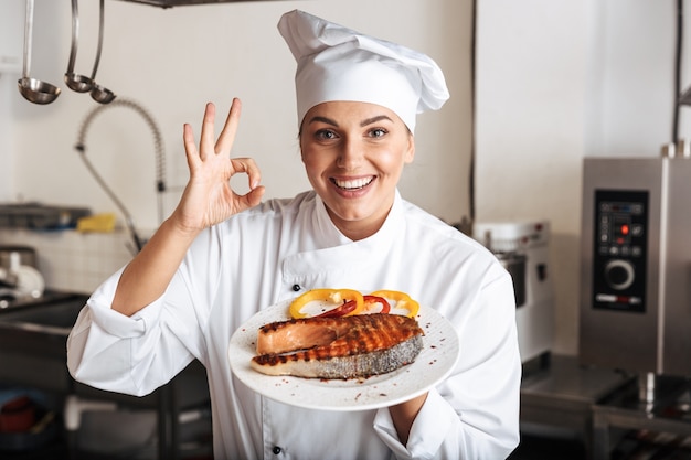 Imagem da chef sorridente, vestindo um uniforme branco, segurando um prato com peixe grelhado na cozinha do restaurante