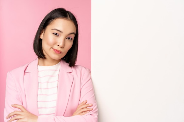 Imagem da jovem empresária asiática bem sucedida senhora corporativa de terno em pé perto do anúncio de informações de parede branca a bordo de braços cruzados e sorrindo como fundo rosa profissional