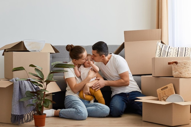 Imagem da jovem esposa e marido posando em sua nova casa no dia da mudança da família com a filha infantil se mudando para um novo apartamento sentado no chão e beijando seu bebê