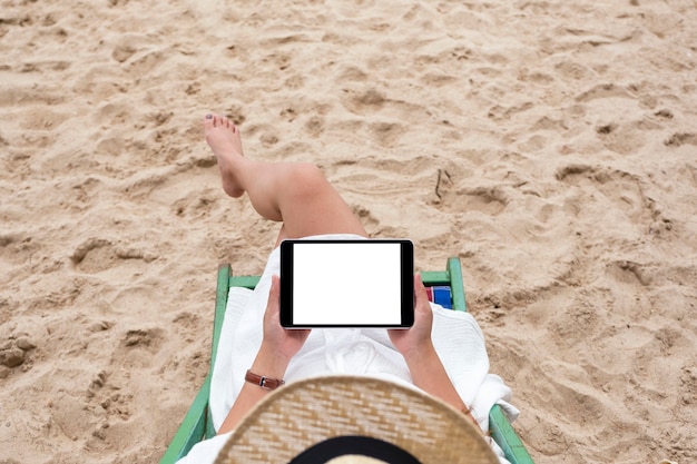 Imagem da maquete de vista superior de uma mulher segurando e usando um tablet pc preto com uma tela em branco enquanto está deitada em uma cadeira de praia