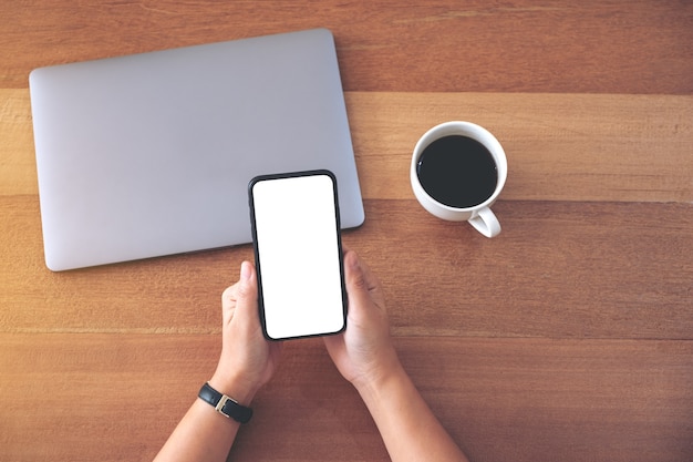 Imagem da maquete de vista superior de uma mulher segurando e usando um telefone celular preto com uma tela de desktop em branco com uma xícara de café e um computador laptop na mesa de madeira