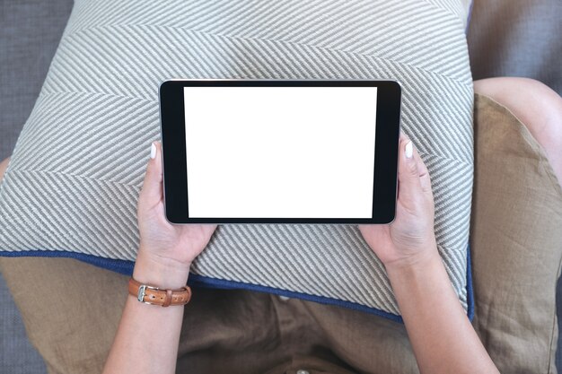 Imagem da maquete de vista superior de uma mulher segurando um tablet pc preto com uma tela em branco na área de trabalho horizontalmente enquanto está sentada na sala de estar sentindo-se relaxada