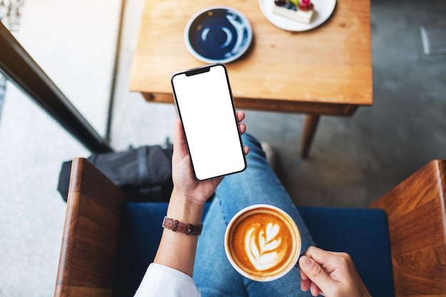 Imagem da maquete de vista superior de uma mulher segurando um telefone celular com uma tela em branco enquanto toma café no café