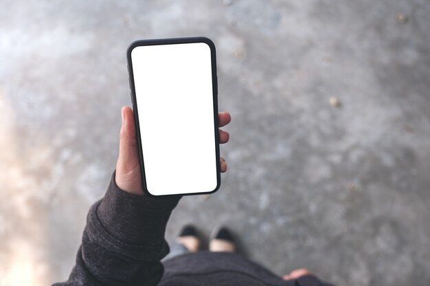 Imagem da maquete de vista superior de uma mulher segurando um telefone celular preto com uma tela em branco enquanto está de pé no chão