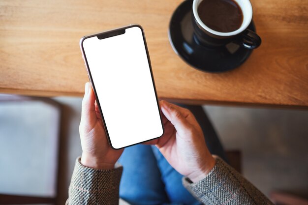 Imagem da maquete de vista superior de uma mulher segurando um telefone celular preto com uma tela em branco enquanto está sentada no café