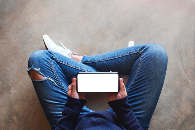 Imagem da maquete de vista superior de uma mulher segurando um telefone celular preto com uma tela em branco enquanto está sentada no chão