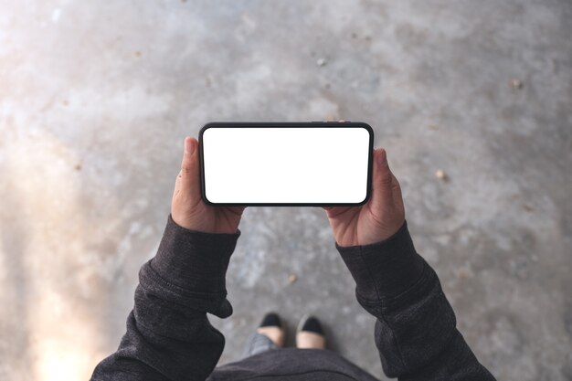 Imagem da maquete de vista superior de uma mulher segurando um telefone celular preto com uma tela em branco horizontalmente em pé no chão de concreto