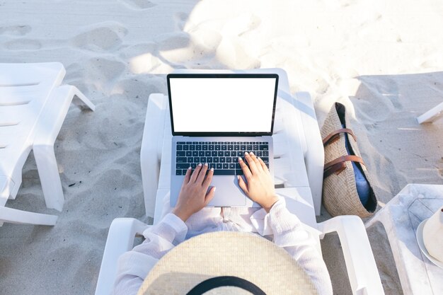 Imagem da maquete de vista superior de uma mulher usando e digitando em um laptop com uma tela em branco enquanto se deita em uma cadeira de praia na praia