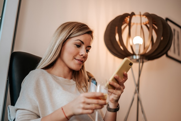 Imagem da mulher loura que aprecia o vidro do vinho branco ao usar o smartphone.