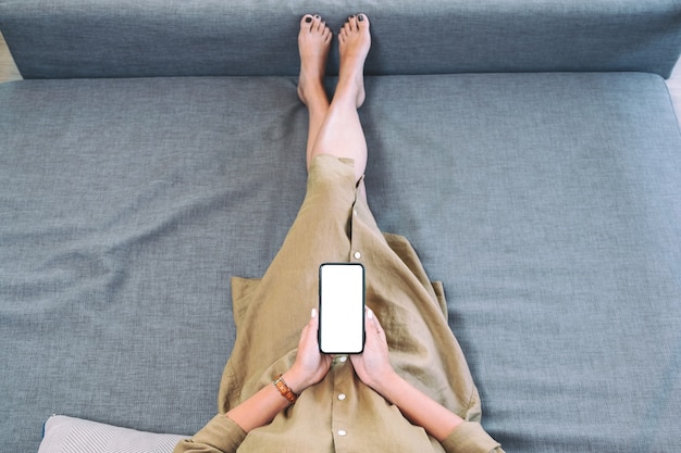 Imagem da vista superior de uma mulher segurando um telefone celular preto com uma tela em branco enquanto se deita na sala de estar sentindo-se relaxada