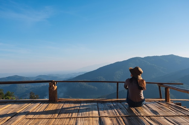 Imagem da vista traseira de um viajante feminino sentado na varanda de madeira e olhando para uma bela vista da montanha