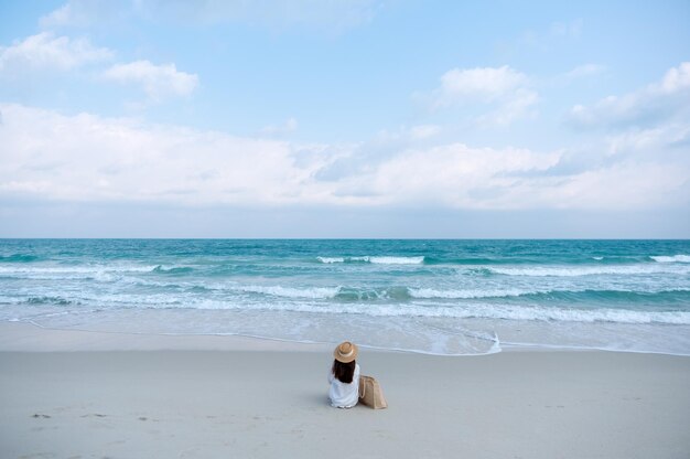 Imagem da vista traseira de uma mulher com chapéu e bolsa sentada na praia com fundo de céu azul