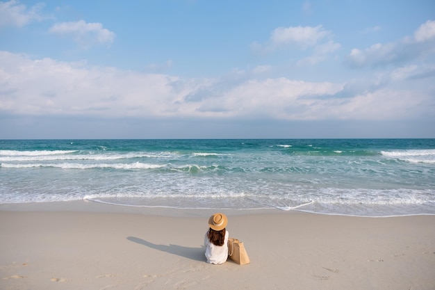 Imagem da vista traseira de uma mulher com chapéu e bolsa sentada na praia com fundo de céu azul