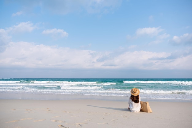 Imagem da vista traseira de uma mulher com chapéu e bolsa sentada na praia com fundo de céu azul