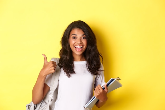 Imagem de alegre aluna afro-americana, segurando cadernos para as aulas e uma mochila, mostrando o polegar para cima, como estudar, em pé sobre um fundo amarelo