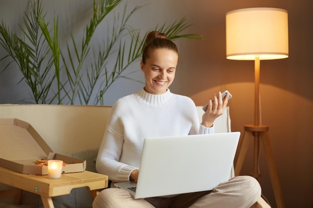 Imagem de alegre atraente jovem adulta vestindo suéter branco sentado no sofá e trabalhando no laptop na sala de estar em casa olhando para a tela do pc e sorrindo