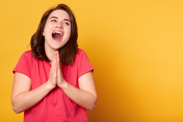 Imagem de alegre caucasiana feminina vestida camiseta casual vermelha, mantém as mãos em gesto de oração, tem feliz expressão facial, seu sonho se torna realidade, fica contra amarelo